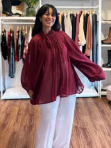 Blusa plisada de color bordó con diseño amplio y fluido. Presenta cuello alto con lazo para anudar, mangas largas con gran volumen y una textura plisada en toda la prenda.