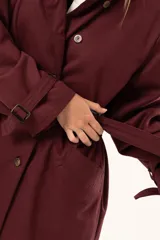 Trench de corte holgado color bordó, con cuello camisero, cierre frontal con botones y cinturón ajustable con hebilla. Presenta mangas largas con detalles de correas en los puños y bolsillos laterales.