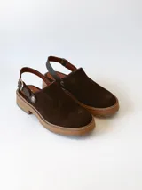 Zueco de cuero gamuzado color chocolate, con diseño de punta cerrada y tira ajustable con hebilla metálica en el talón. Presenta una suela de plataforma dentada en tono marrón claro.