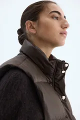Chaleco acolchado tipo puffer color marrón oscuro, con cuello elevado, cierre frontal oculto por solapa con botones a presión y bolsillos tipo parche en el frente.