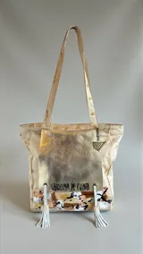 Cartera tipo tote de cuero color natural con bolsillo frontal con solapa metalizada en tono dorado, detalles pintados a mano en la base, flecos decorativos y doble asa de hombro.