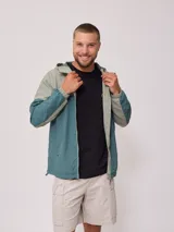 Campera rompevientos con cierre frontal, confeccionada en nylon con diseño de bloques de color en tonos verde azulado y gris claro. Cuenta con capucha, puños elásticos y cordón de ajuste en el ruedo.