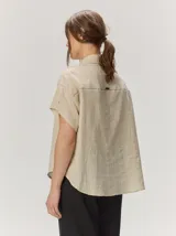 Camisa de manga corta color beige con sutil diseño a rayas verticales, cuello camisero con detalle de botones decorativos en la solapa y cierre frontal oculto.