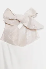 Vestido corto de tela brillante color blanco perla, con cuello halter que se anuda en la parte superior, escote drapeado y bajo con volantes superpuestos.