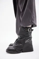 Botas de caña media confeccionadas en cuero vegano color negro, con diseño de solapa frontal y detalle de hebilla metálica en la parte superior.