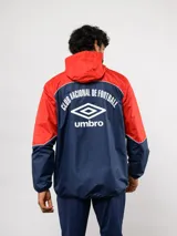 Campera de lluvia Umbro del Club Nacional de Football, con diseño de bloques de color en rojo y azul marino. Presenta capucha, cierre frontal, puños elásticos y detalles de piping blanco. Incluye logo de Umbro y escudo del club en el pecho.