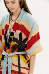 Kimono largo de lino con estampado vibrante que simula un paisaje de atardecer con palmeras, en franjas horizontales de colores cálidos y verdes. Posee mangas amplias y lazo para ajustar en la cintura.