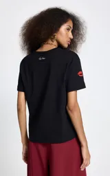 Remera negra de algodón con cuello redondo y mangas cortas. Presenta un bordado frontal con la frase 'L'amour est déclaré' acompañada de un diseño de labios rojos, y un detalle bordado adicional en la manga derecha.