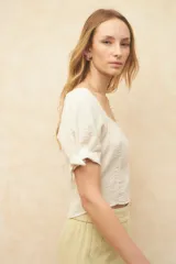 Top corto color blanco roto, con escote cuadrado, manga corta abullonada con lazos ajustables en el borde y textura arrugada en el tejido.