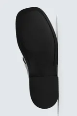 Zueco destalonado de cuero negro con hebilla metálica ajustable en el empeine, plantilla de cuero y suela plana con pequeño tacón.