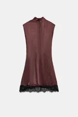 Vestido corto de cuello subido y sin mangas, confeccionado en tejido satinado. Presenta un detalle de fruncido lateral con abertura frontal y aplicación de encaje en el bajo.