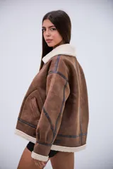Campera tipo gamulán de corte oversize, confeccionada en cuero sintético marrón con efecto desgastado y forro interno de corderito sintético color crema. Presenta cuello solapa, cierre frontal con cremallera, detalles de tiras con hebillas en el cuello y en los laterales, y costuras reforzadas con bandas de cuero oscuro.