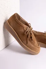 Zapato tipo mocasín acordonado de gamuza color marrón oscuro, con suela de goma color caramelo y detalle de dije metálico dorado en forma de bota colgando de los cordones.