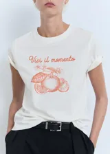 Camiseta blanca de manga corta y cuello redondo, confeccionada en algodón. Presenta un estampado frontal con la frase "Vivi il momento" y un gráfico de naranjas en color rojo.