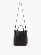 Tote bag Jazmin Chebar de cuero vacuno negro con textura grabada. Presenta un diseño compacto con doble asa superior y un tag colgante con un aplique metálico en forma de labios.
