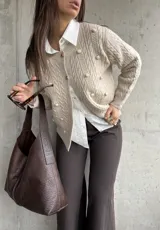 Cardigan de punto color beige con diseño de trenzas y pompones decorativos. Presenta cuello redondo, cierre frontal con botones y mangas largas.
