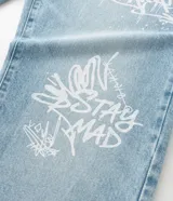 Pantalón de jean corte baggy, color celeste claro con efecto desgastado y estampado de lettering estilo grafiti en color blanco. Presenta cintura con trabillas, cierre frontal con botón y cremallera, y bolsillos laterales y traseros.