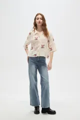 Camisa de algodón color crudo con diseño de paneles unidos por cintas caladas. Presenta bordados florales en cada panel, cuello camisero, cierre frontal con botones y mangas tres cuartos.