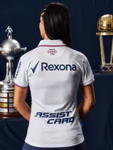 Camiseta de fútbol Umbro del Club Nacional de Football, modelo Home 2026 para mujer. Diseño en color blanco con textura jacquard que presenta siluetas de copas históricas en toda la prenda. Cuenta con cuello en V, detalles en azul y rojo en los bordes de las mangas y el cuello, logo de Umbro, escudo del club y sponsor Antel en el frente.