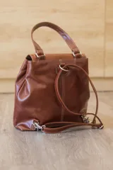 Mochila de cuero vacuno marrón con acabado pulido, solapa en forma de V y costuras decorativas. Cuenta con cremalleras laterales y asas ajustables.