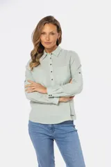 Camisa blanca de manga larga con cuello y tapeta de medio botón. Presenta dos bolsillos frontales con tapa y botones metálicos. Las mangas tienen presillas para sujetar el puño enrollado.