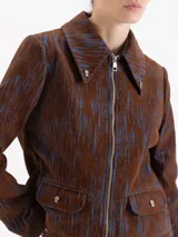 Chaqueta de corte recto con cierre frontal, cuello camisero amplio y bolsillos delanteros con solapa y botones metálicos. Presenta un diseño estampado con líneas verticales en tonos marrón y azul.