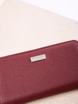 Billetera rectangular de cuero sintético con textura granulada, color bordó. Cuenta con cierre perimetral metálico y placa metálica con el logo de la marca en el frente.
