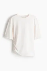 Remera de algodón color blanco crudo, con cuello redondo, mangas cortas y detalle de fruncido lateral que genera un efecto drapeado y un bajo asimétrico.
