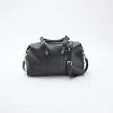 Bolso de mano estilo bowling en color negro, confeccionado en material texturizado. Presenta doble asa superior con detalles de hebillas decorativas, cierre principal con cremallera y correa larga ajustable y extraíble para llevar al hombro o cruzado. Incluye herrajes metálicos en tono dorado.