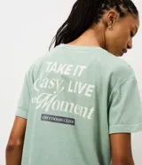 Remera de algodón con calce oversized, cuello redondo y mangas cortas. Presenta un diseño minimalista con un pequeño estampado de texto en el pecho y un acabado lavado en color verde menta.