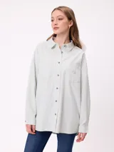 Camisa de corte oversize con diseño de rayas verticales en tonos verde claro y blanco. Presenta cuello clásico, cierre frontal con botones y un bolsillo en el pecho con detalle bordado de la marca.