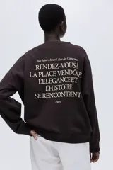 Buzo de tejido de punto color marrón oscuro, con cuello redondo, hombros caídos y puños y bajo acanalados. Presenta un pequeño texto estampado en color blanco en el pecho que dice "RUE DES CAPUCINES Paris, 1991".