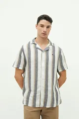 Camisa de manga corta con cuello cubano, confeccionada en una mezcla de lino y algodón con textura. Presenta un diseño a rayas verticales en tonos beige y celeste sobre fondo blanco, con cierre frontal de botones y logo bordado en el pecho.