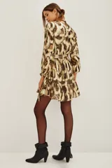 Vestido corto de estilo wrap con estampado abstracto en tonos dorados y marrones con acabado metalizado. Presenta escote en V cruzado, mangas largas con puños elásticos, lazo ajustable en la cintura y falda con volado en el ruedo.