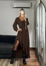 Vestido largo color marrón, con escote en V, mangas largas con puños y detalle de encaje en el pecho. Presenta un diseño asimétrico con volados en el ruedo y una abertura frontal.