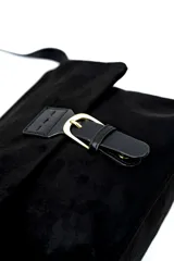 Cartera de gamuza y cuero sintético negro, con solapa frontal, tira ajustable para el hombro y detalle de hebilla metálica dorada en el cierre.