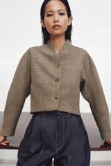 Chaqueta corta de corte estructurado confeccionada en tejido con mezcla de lana. Presenta cuello en pico, cierre frontal con botones y mangas largas. El diseño incluye bolsillos delanteros y un forro interior a contraste.