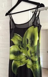 Vestido largo de microtul negro con estampado de flores grandes en tonos verdes. Presenta breteles finos, escote redondo y diseño ajustado al cuerpo con transparencias.