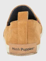 Alpargata Hush Puppies de gamuza color mostaza, con suela de yute trenzado y base de goma. Presenta un detalle elástico lateral con diseño multicolor y forro interior de algodón orgánico.