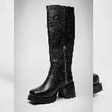 Bota de caña alta confeccionada en cuero vacuno, con diseño de punta cuadrada y plataforma. Presenta un taco cuadrado de madera, cierre lateral interno y una tira decorativa con herraje metálico circular en el tobillo.