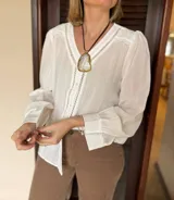 Camisa blanca de tela liviana con textura sutil, presenta detalles calados verticales en el delantero y volados delicados en los hombros. Posee mangas largas con puños y cierre frontal mediante botones.