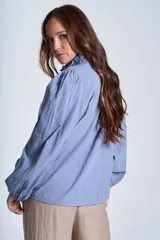 Camisa de color azul con cuello alto decorado con volados y lazo de ajuste. Presenta tablas decorativas en los hombros, cierre frontal con botones de nácar y mangas largas con puños elásticos que generan un efecto abullonado.