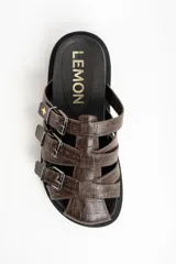 Zueco tipo pescador de cuero marrón con textura cocodrilo. Presenta tres tiras ajustables con hebillas metálicas plateadas sobre el empeine. Tiene una suela negra gruesa y dentada.