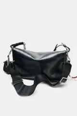 Bolso de hombro tipo baguette en color negro, con acabado brillante y estructura rectangular. Cuenta con una correa combinada de cadena metálica plateada y cuero sintético, y un pequeño monedero extraíble unido a un lateral. Presenta detalles de cordones decorativos en el otro lateral y cierre mediante solapa con imán.