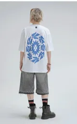 Remera blanca de corte holgado con estampa central en color azul que combina motivos geométricos tipo chakana con figuras de aves dispuestas en círculo.