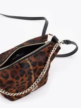 Bandolera de tamaño pequeño confeccionada en pelo vacuno con estampado animal print de leopardo y detalles en cuero vacuno flor. Presenta una cadena metálica decorativa en el frente, aplique metálico con logo JCH y correa de cuero ajustable.