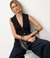 Chaleco negro de tejido punto, sin mangas, con cuello solapa y cierre frontal de dos botones dorados. El corte es acinturado y alargado.