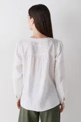 Blusa de lino color blanco, con cuello redondo, cierre frontal con botones de madera y dos bolsillos delanteros. Presenta mangas largas con puños abotonados y costuras a la vista en contraste.