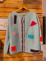 Saco de lana Merino tejido a mano con dos agujas, en color celeste claro, con cierre frontal de botones de madera y parches geométricos de diversos colores aplicados en el frente y las mangas.