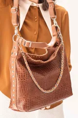 Cartera tipo hobo de cuero vacuno con textura grabada efecto croco en color tabaco. Presenta detalles de costuras artesanales, laterales con apliques metálicos y una cadena decorativa frontal. Incluye una correa corta para hombro y una correa larga extensible y desmontable, ambas con herrajes metálicos.